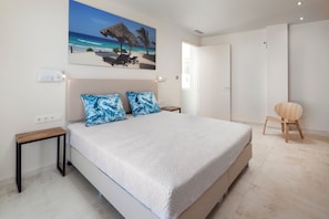 A bedroom with a bed, a nightstand, a chair, a painting of a beach scene, and a door.
