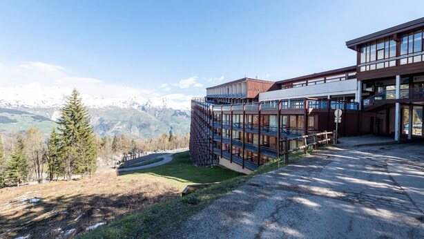 Um edifício moderno com fachada de vidro, área de estacionamento e vista para montanhas nevadas.