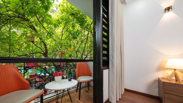 A balcony with a view of green trees, a small table with chairs, and a nightstand with a lamp. - Hanoi Liliane Hotel and Travel (Hanoi)