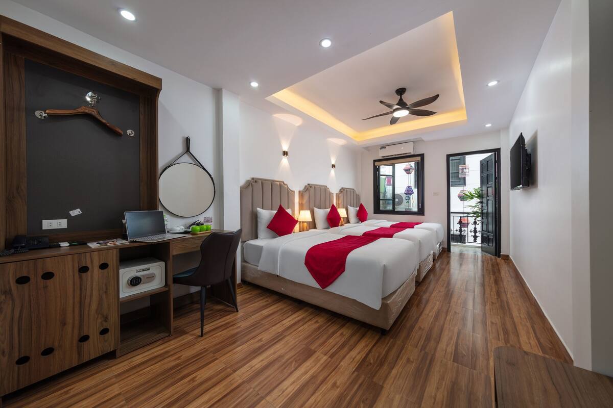 Een moderne hotelkamer met een houten bureau, een bed met rode kussens, een plafondventilator en een televisie aan de muur.