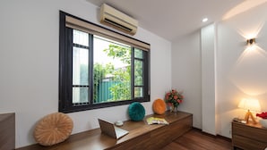 A modern room with a wooden desk, a laptop, a window with blinds, a bedside table with a lamp, and a wall-mounted air conditioner. - Hanoi Liliane Hotel and Travel (Hanoi)