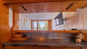 L’intérieur d’une cabane en bois comprend un comptoir de cuisine, des armoires en bois, une fenêtre donnant sur des arbres et des montagnes, ainsi qu’un téléviseur fixé au mur.