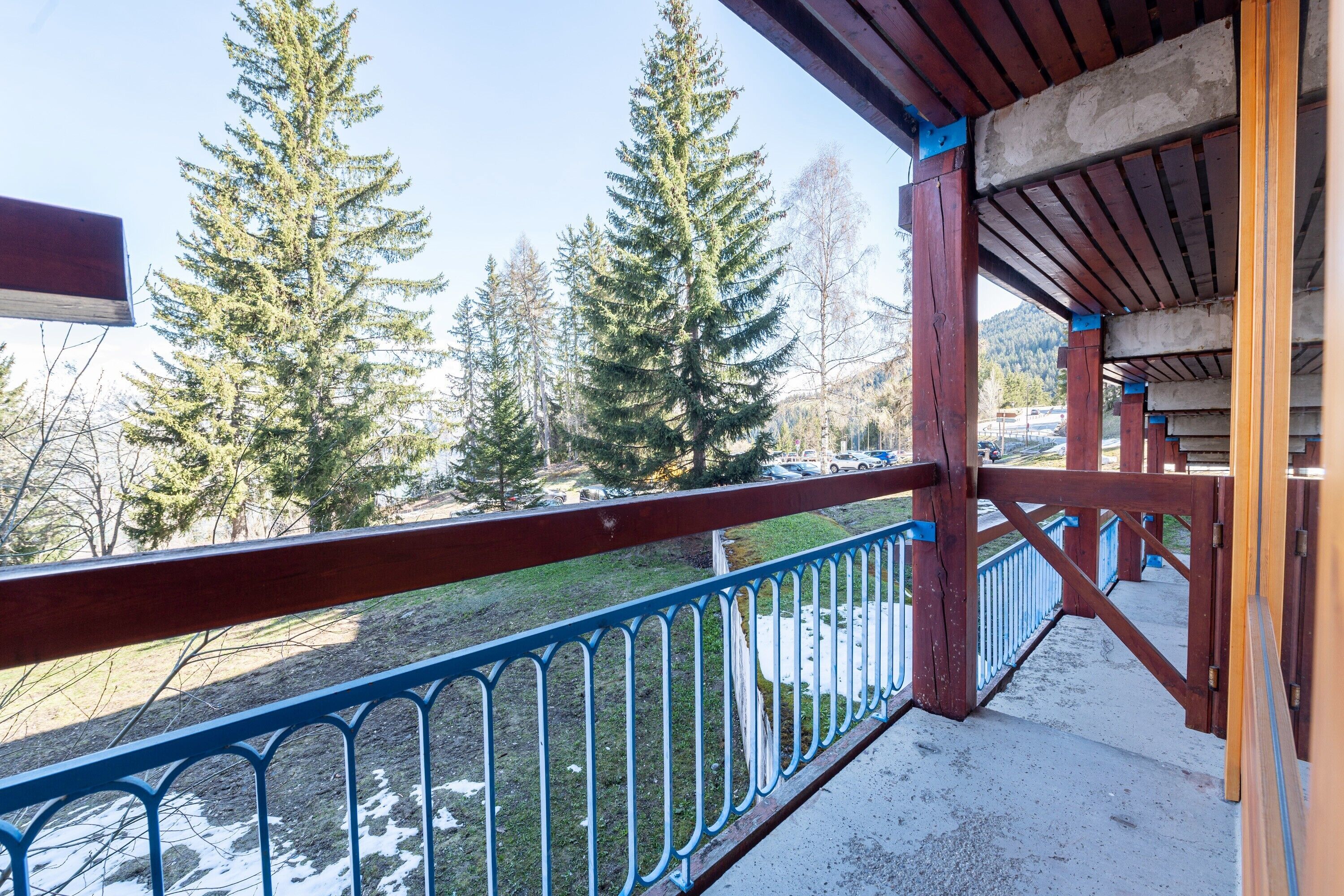 A wooden balcony with a metal railing overlooking a snowy landscape with trees.