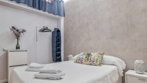 Een netjes opgemaakt bed met witte lakens, een nachtkastje met een lamp, een kledingrek en een wandplank.