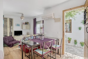 Een moderne woonkamer met een glazen eettafel, een paars tafelkleed en bijpassende stoelen. Er is een televisie op een kast, een bank en een potplant op de vensterbank.