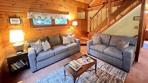 Interior acolhedor de uma cabana de madeira com dois sofás cinzas, uma mesa de centro em madeira e uma escada.