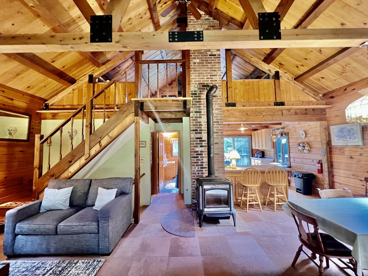 A cozy cabin interior with a wooden staircase, a sofa, a table, and a wood-burning stove.