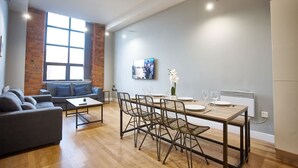 A modern living room with a sofa, coffee table, dining table with chairs, a TV mounted on the wall, and a large window. - The Onyx Suite Summar (Manchester)