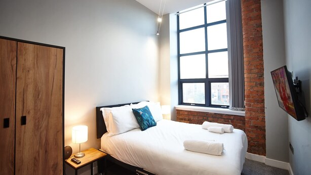 A hotel room with a bed, a nightstand, a lamp, a window with a view, and a brick wall. - The Onyx Suite Summar (Manchester)