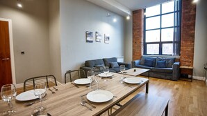 A dining area with a long wooden table set for four, a grey sofa, and a large window with a view. - The Onyx Suite Summar (Manchester)