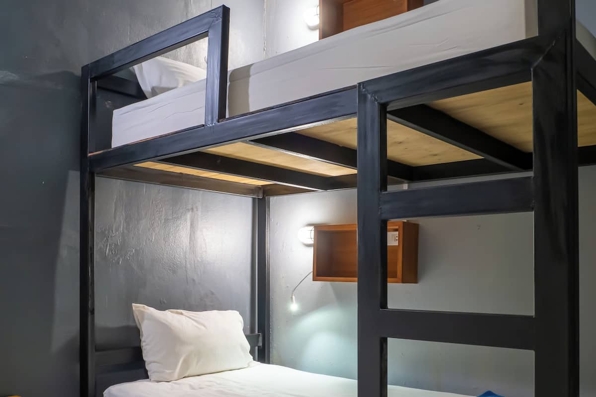 A bunk bed with a white mattress and pillow, a wooden shelf above, and a small wooden nightstand beside it.