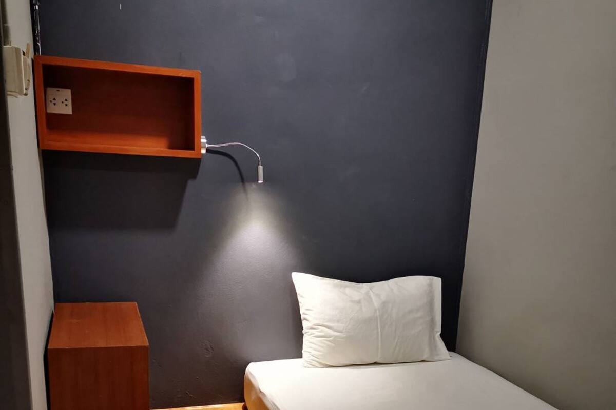 A small, compact room with a single bed, a wooden nightstand, a shelf with a mirror, and a wall-mounted light.