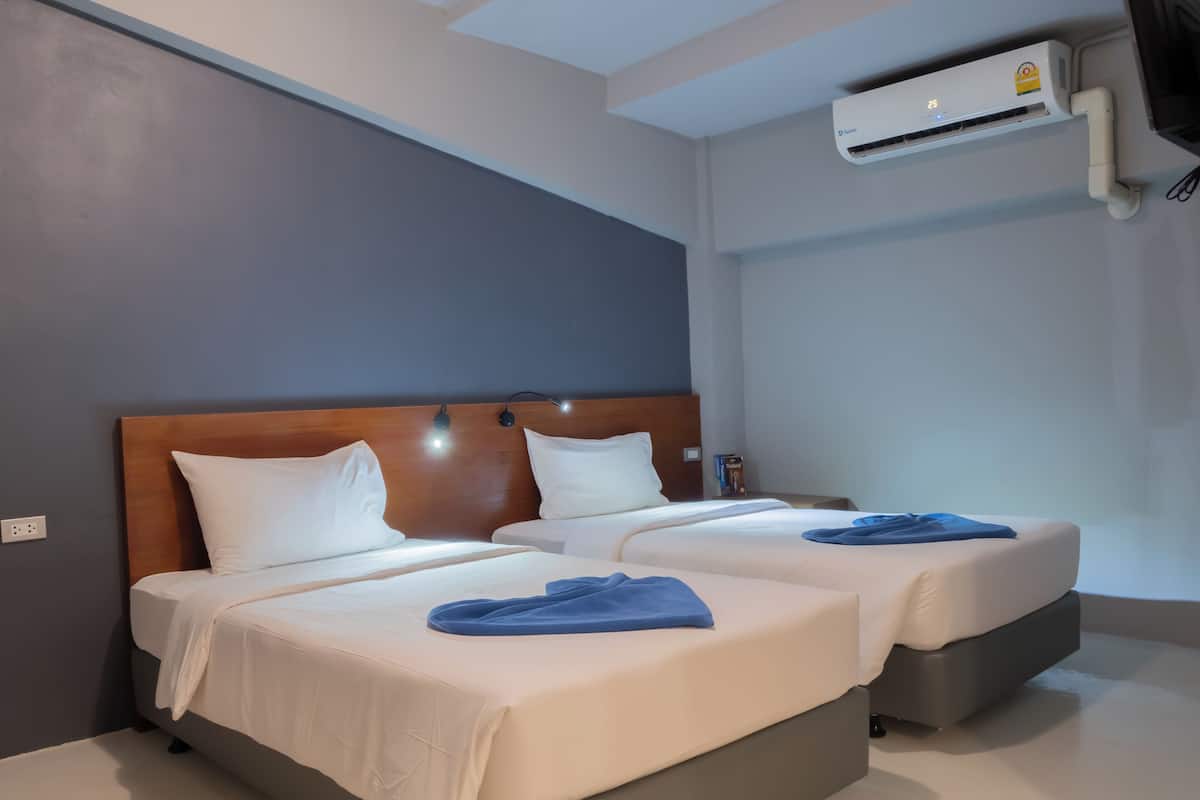 A hotel room with two beds, a wooden headboard, white bedding, a blue towel, an air conditioning unit, and a nightstand with a remote control.