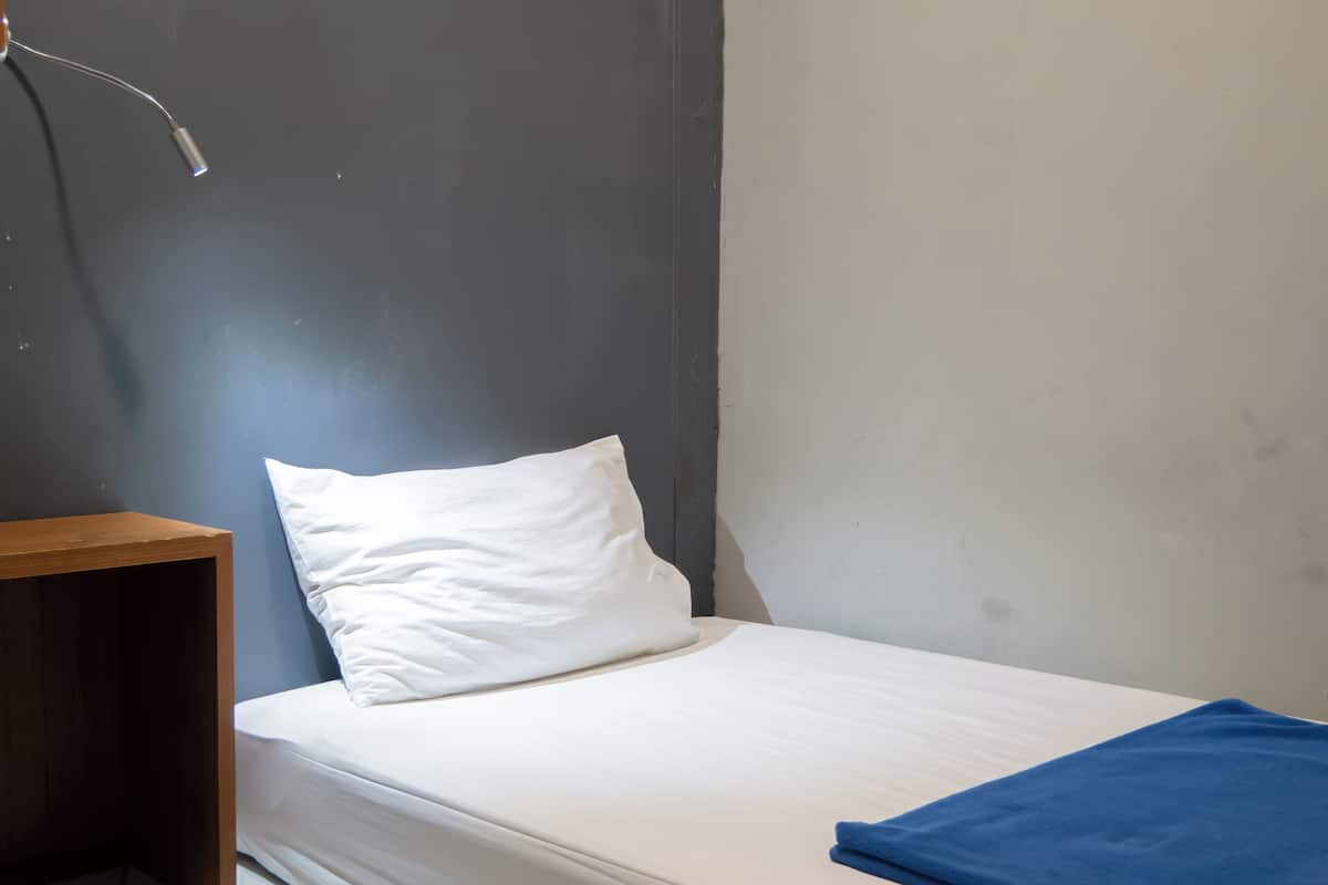 A small, neatly made bed with a white mattress and a single white pillow. There is a blue folded blanket on the bed. A wooden nightstand is positioned to the left of the bed. The room has a simple, modern design with a grey wall and a white floor.