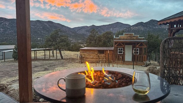 Eine Terrasse mit Feuerstelle, einem Tisch und einem Weinglas. Im Hintergrund ein Holzhäuschen und Berge.