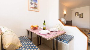 Un coin repas avec une table dressée pour deux personnes, un saladier de fruits, une bouteille de vin et un tableau encadré au mur.
