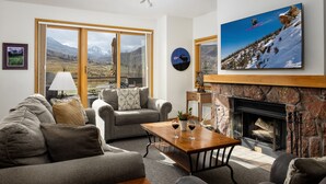 Un salon doté d’une cheminée, d’un ensemble de canapés, d’une table basse et d’une grande fenêtre donnant sur une vue imprenable sur les montagnes.