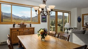 Un espace repas comprenant une table en bois, des chaises, un lustre et une vue sur les montagnes, visible à travers de grandes fenêtres.