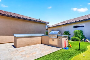 An outdoor barbecue area with a grill and a built-in cabinet, surrounded by a paved patio and a well-maintained lawn.