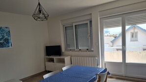 Una habitación con una mesa de comedor, sillas, un televisor en un mueble bajo, una ventana con persianas y vistas a edificios.