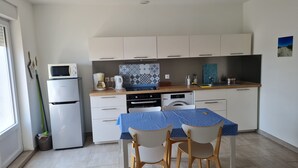 Cocina pequeña con armarios blancos, microondas, cafetera y lavavajillas. Cuenta con una mesa de comedor con sillas y mantel azul.