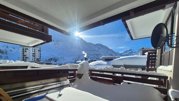 Ein mit Schnee bedeckter Außenbereich mit Sitzgelegenheiten und Blick auf verschneite Berge sowie eine Seilbahn im Hintergrund.