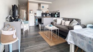 Een ruimtelijk ingerichte leefruimte met kitchenette, een bank, een eettafel en een televisie.