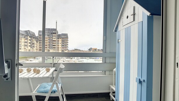 Een kamer met een groot raam, een witte stoel met een blauw kussen, een kleine tafel en een blauw-witte kast.