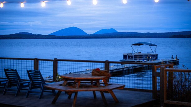 A lakeside deck with a table and chairs, a boat docked at the pier, and mountains in the background. - Sleeps 25! Book all 5 Lawrence`s Lakeside Cabins! (Rockwood)