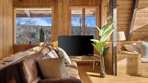 A cozy cabin interior with a wooden ceiling, a leather sofa, a flat-screen TV, and a large window offering a view of a snowy forest. - Cozy Mt. Snow Cabin w/ Fireplace (Wilmington)