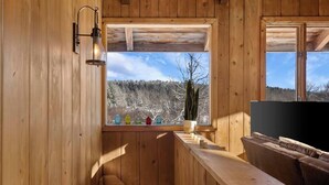 A wooden cabin interior with a wooden bench, a small table, and a window with a view of a snowy forest. - Cozy Mt. Snow Cabin w/ Fireplace (Wilmington)