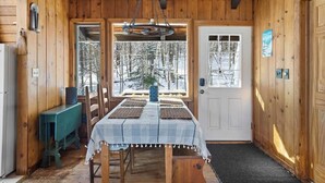 A wooden cabin interior with a dining table, chairs, a hanging light fixture, and a door leading outside. - Cozy Mt. Snow Cabin w/ Fireplace (Wilmington)