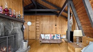 A cozy room with a stone fireplace, wooden walls, a leather sofa, and a wooden table with a lamp. - Cozy Mt. Snow Cabin w/ Fireplace (Wilmington)