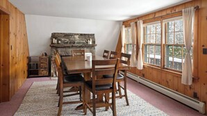 A dining area with a wooden table and chairs, a stone fireplace, and large windows with curtains.