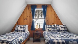 A cozy attic bedroom with two beds, a small table, and a window with curtains.