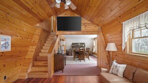A wooden cabin interior with a staircase leading to a dining area, a sofa, and a flat-screen TV mounted on the wall.