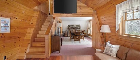A wooden cabin interior with a staircase leading to a dining area, a sofa, and a flat-screen TV mounted on the wall.