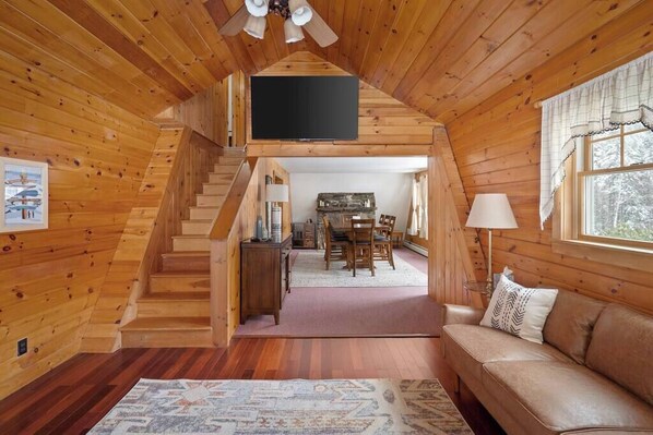 Interior de una cabaña de madera con una escalera que lleva a un comedor, un sofá y un televisor de pantalla plana montado en la pared.