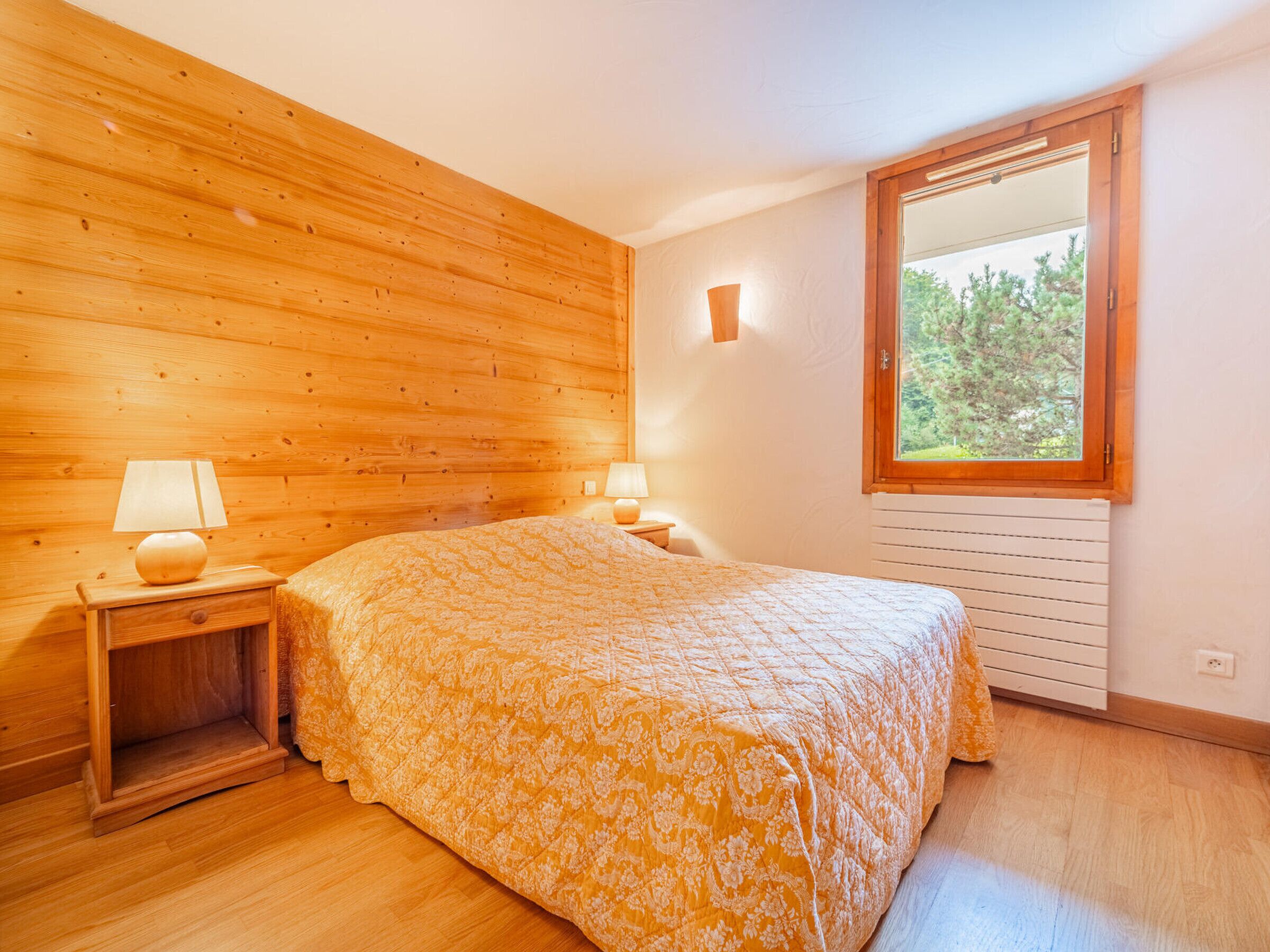 A bedroom with wooden walls, a large bed with a patterned bedspread, two bedside lamps, and a window with a view of trees.