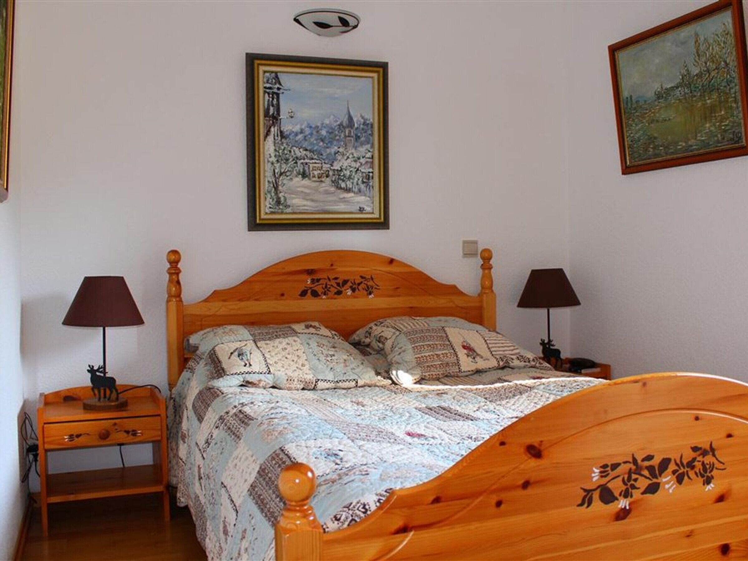 A bedroom with a wooden bed, two bedside tables, a nightstand, two lamps, and two paintings on the walls.