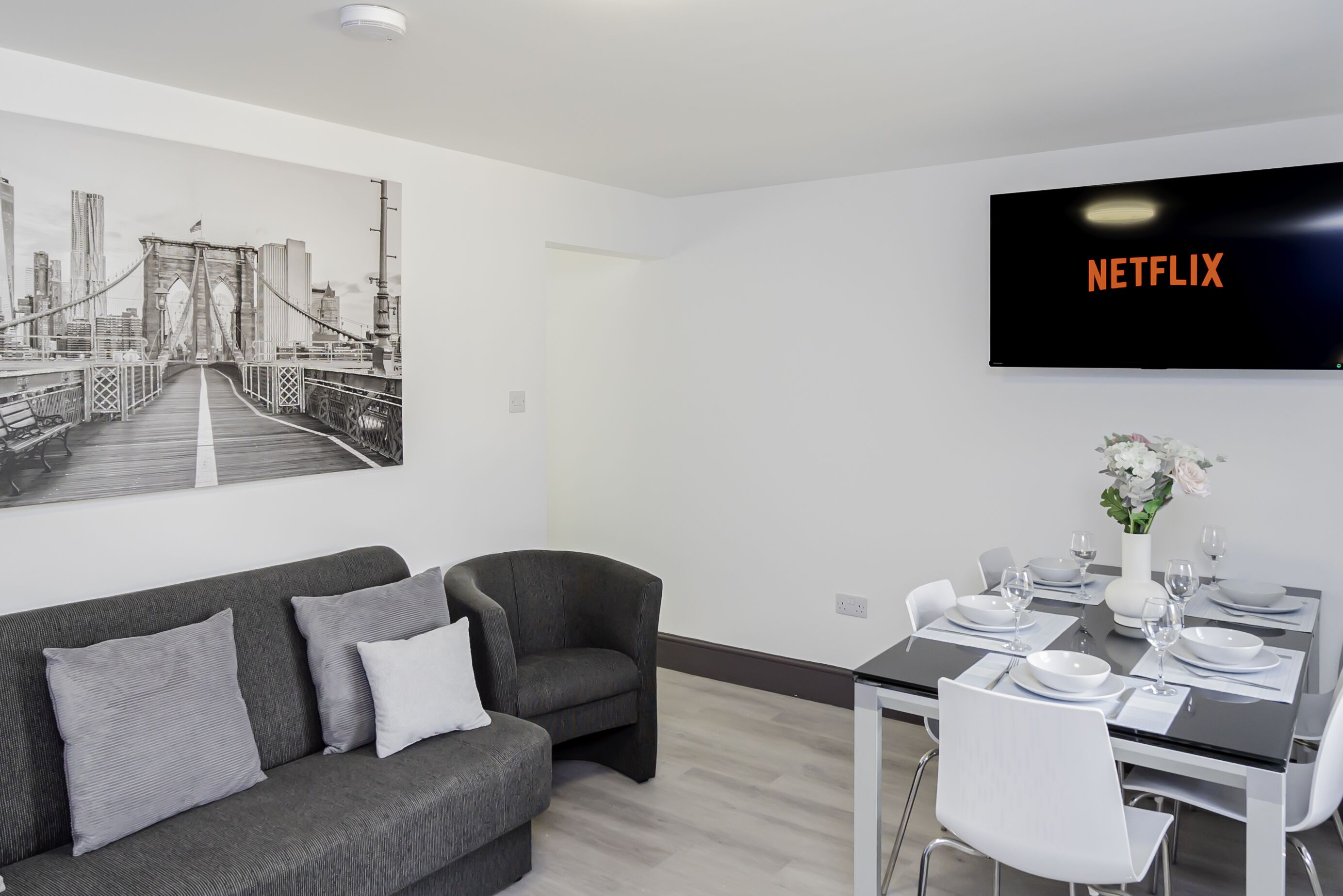 Un salon moderne avec un canapé gris, une table à manger dressée pour quatre personnes et un grand téléviseur fixé au mur, affichant le logo « NETFLIX ».