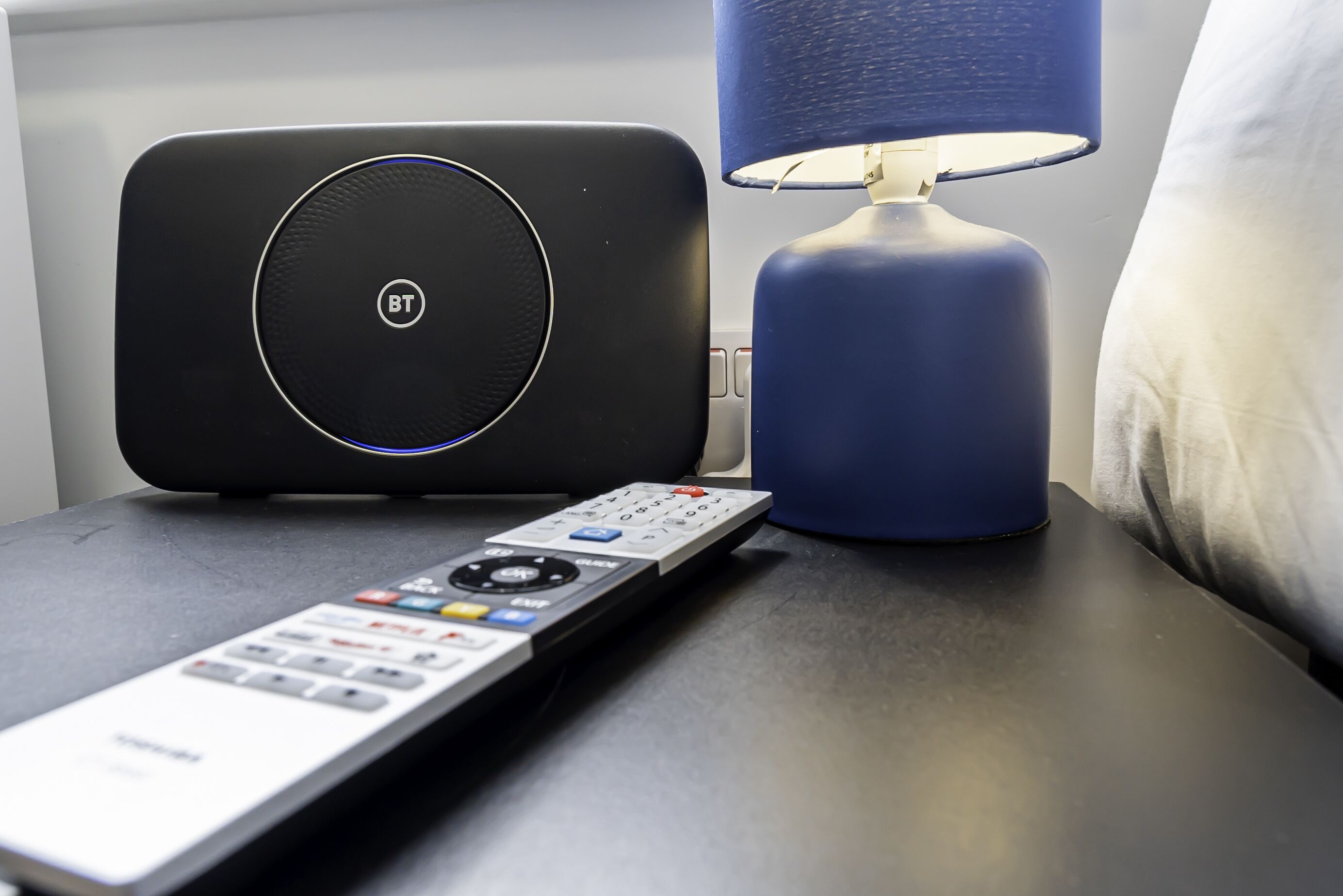 Un control remoto, un altavoz negro y una lámpara azul sobre una mesita de noche.