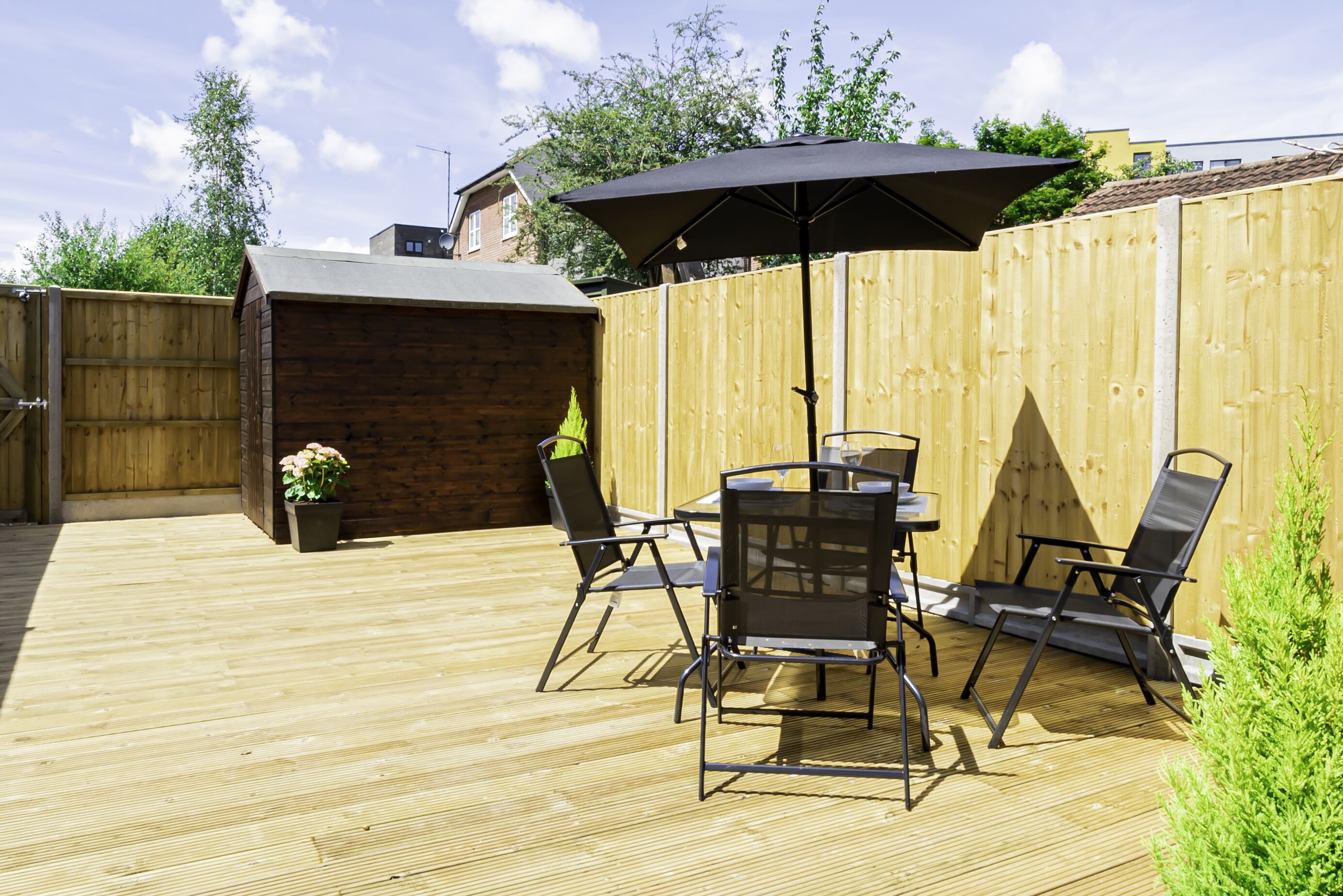 Terraza de madera con una mesa y sillas bajo un gran paraguas, rodeada por una valla de madera.