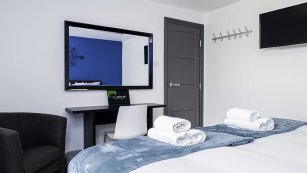 A hotel room with a bed, a desk, a chair, a TV, and a coat rack.