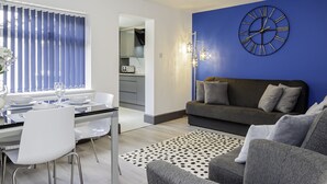 A modern living room with a blue accent wall, a round clock, and a dining area with a glass table and white chairs.