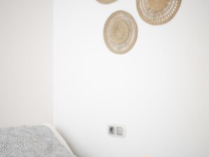 Quarto com parede branca, manta cinza na cama e três tapeçarias trançadas.