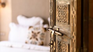 Une porte en bois ouvragée, ornée de sculptures complexes, s’ouvre sur une chambre où se trouve un lit recouvert de draps blancs et agrémenté d’un coussin aux couleurs vives.