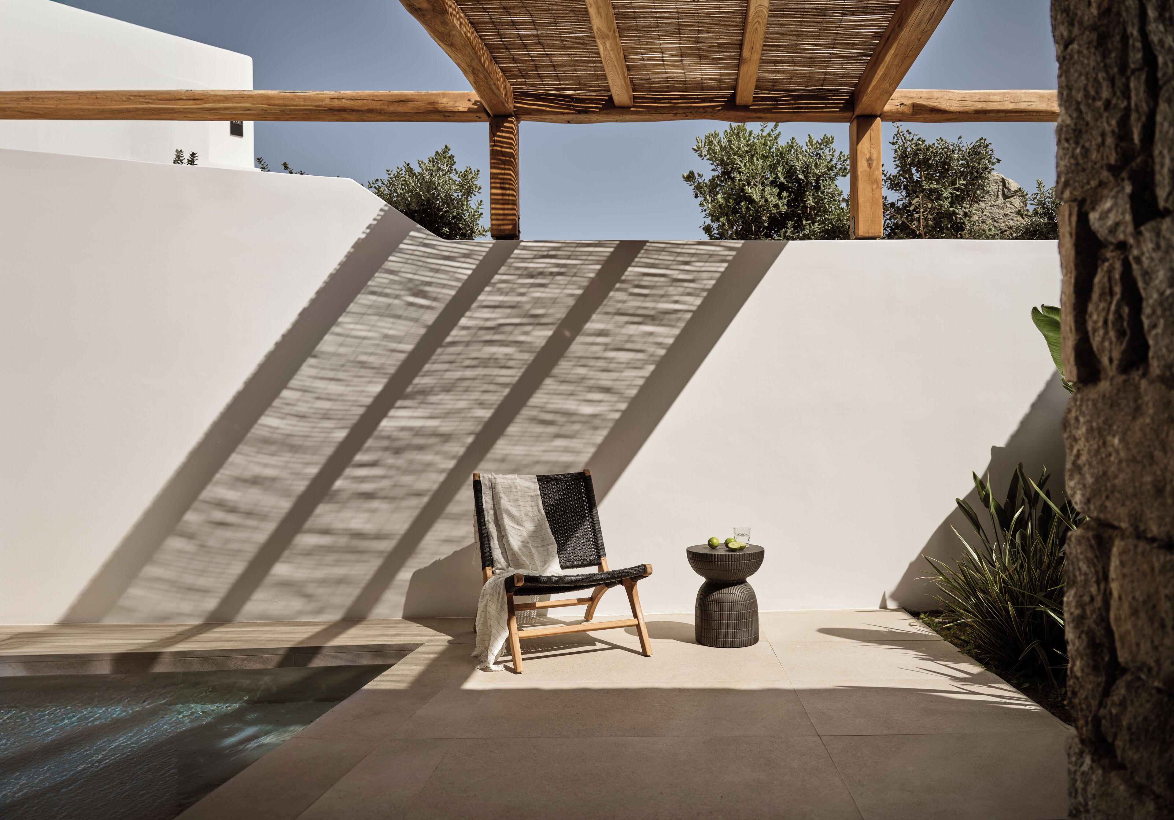 Un espacio al aire libre iluminado por el sol, con una silla de madera, una mesa redonda pequeña y una piscina.