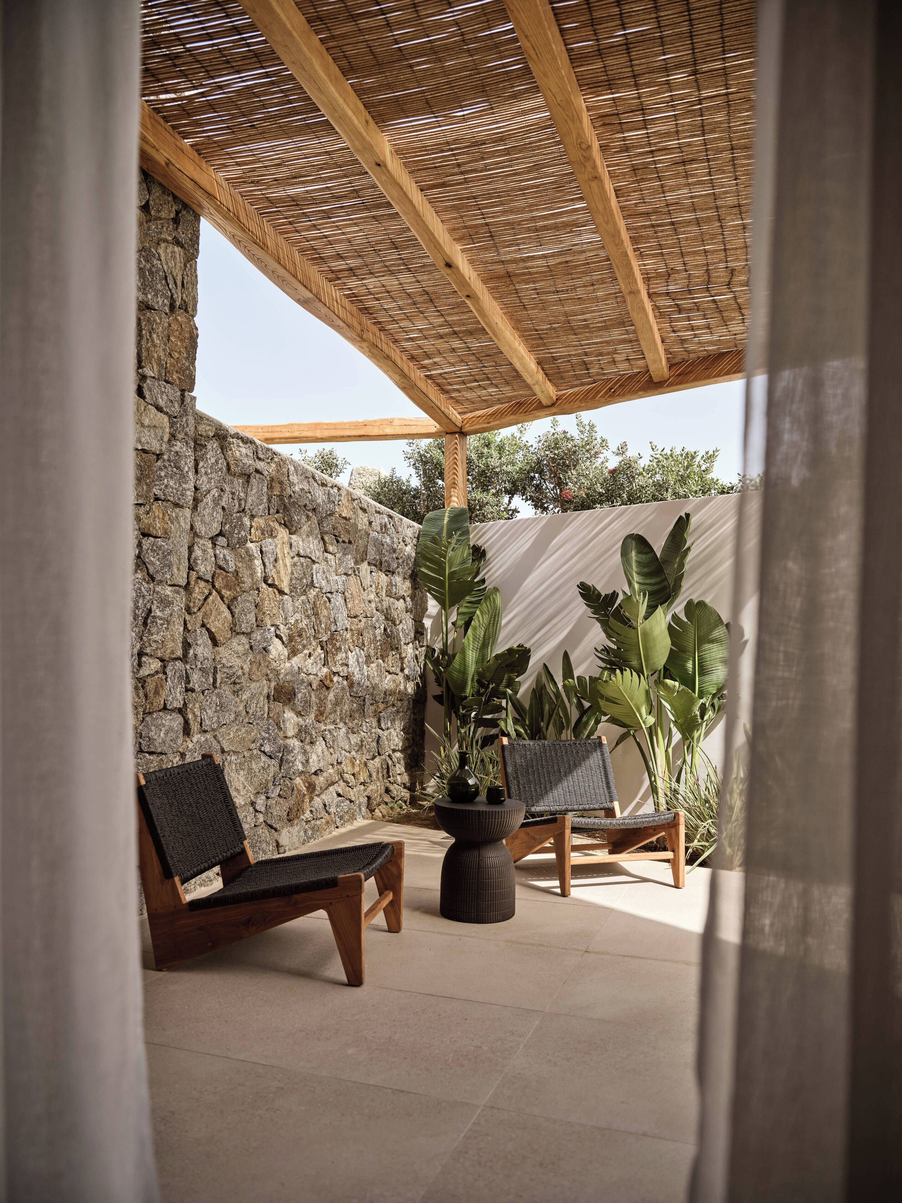 A covered outdoor seating area with stone walls, large potted plants, and wooden furniture.