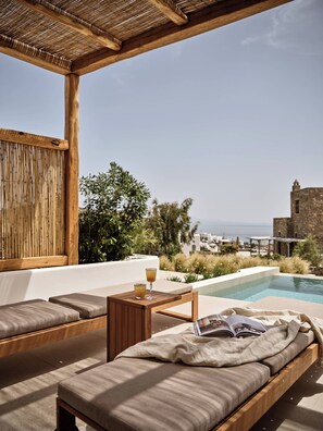 A sunlit outdoor seating area with a pool, wooden furniture, and a view of the sea and a stone building. - Anemelia Hotel Mykonos (Mykonos)